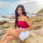 Woman in a red top and white shorts sitting on rocks by the ocean.