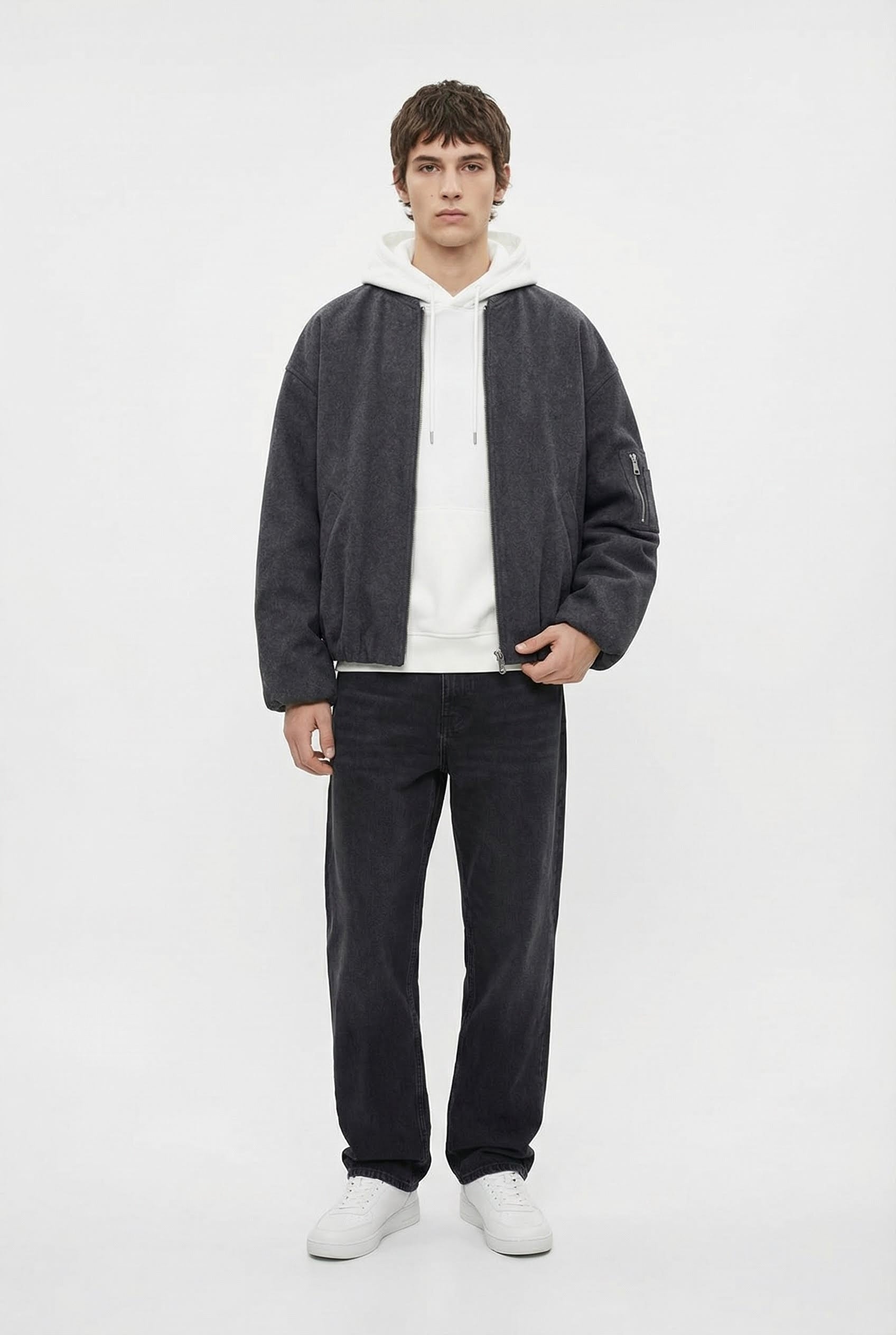 Person wearing a dark gray jacket, white shirt, black pants, and white shoes on a white background